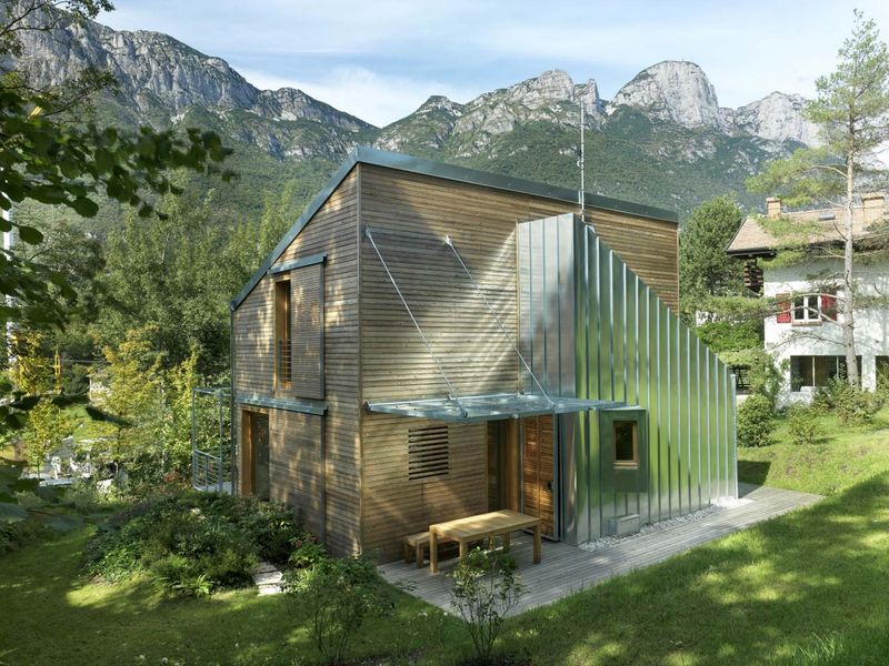 Casa Adhmad di ILLE rivestita in doghe di legno con un volume in lamiera metallica, giardino e pareti di roccia sullo sfondo