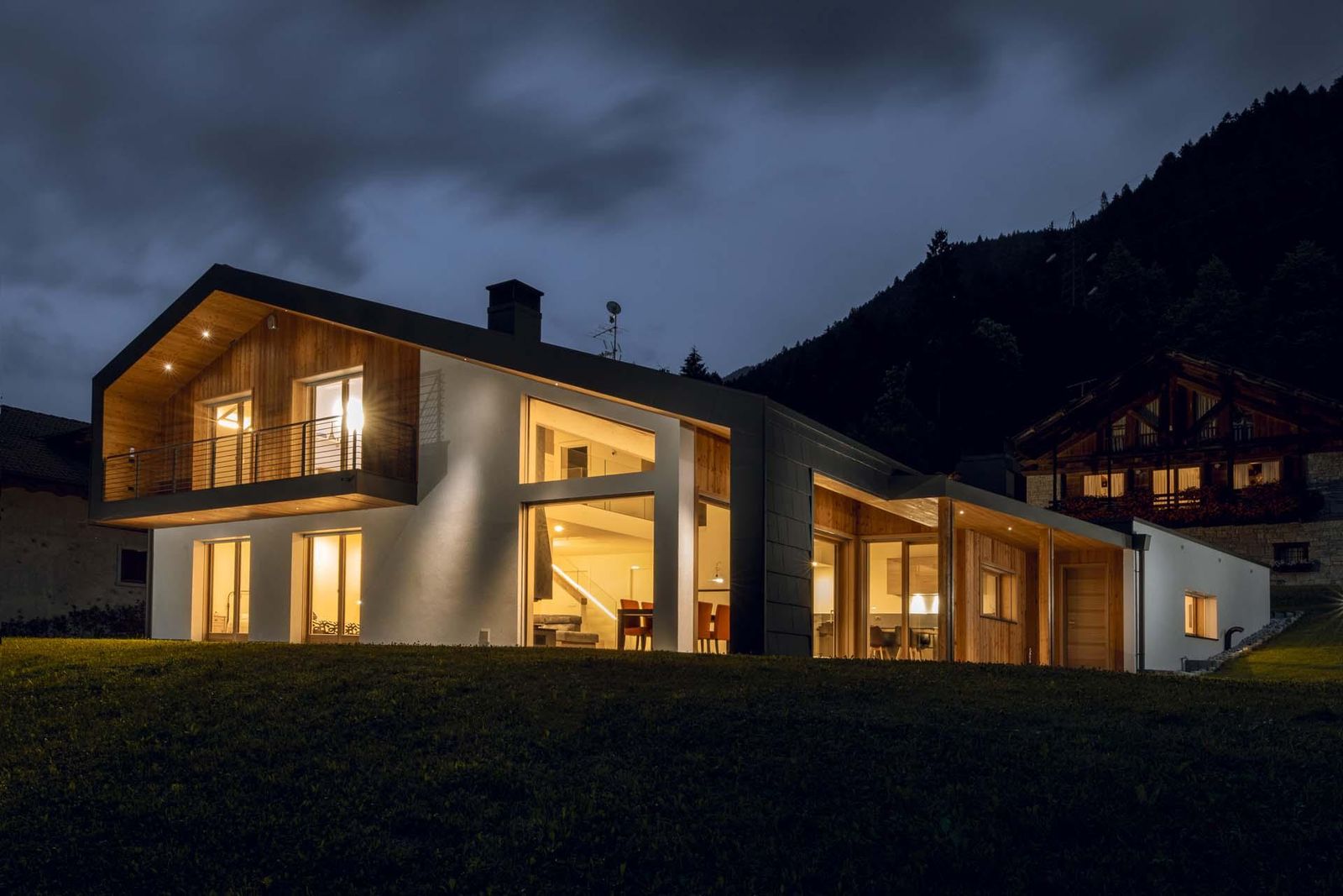 Villa in legno ILLE di notte con grandi vetrate illuminate, tetto a falde scuro e balcone, ai piedi di un versante boscoso
