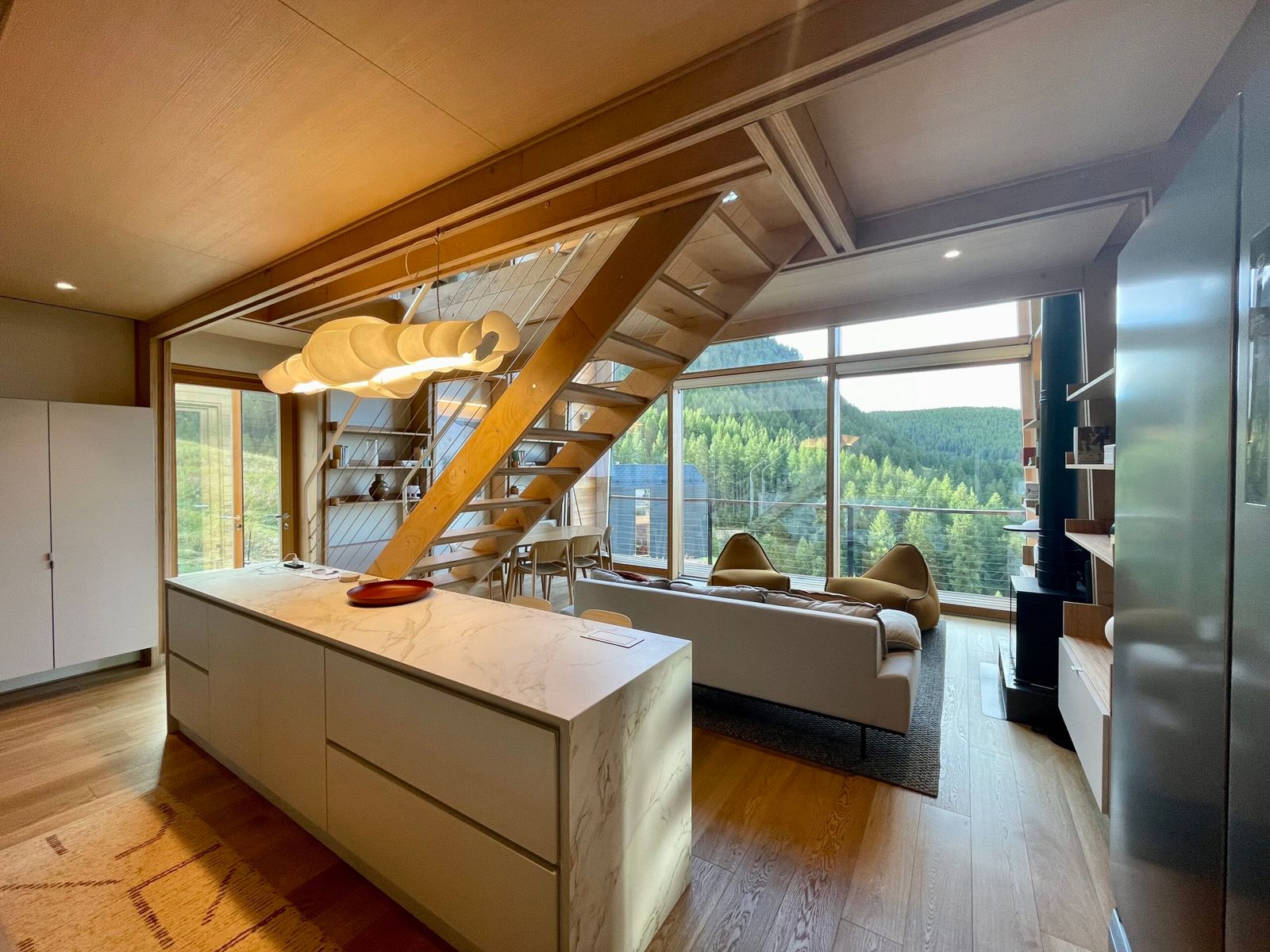 Cucina con isola bianca, scala in legno a vista e soggiorno a doppia altezza davanti a una vetrata sulle montagne