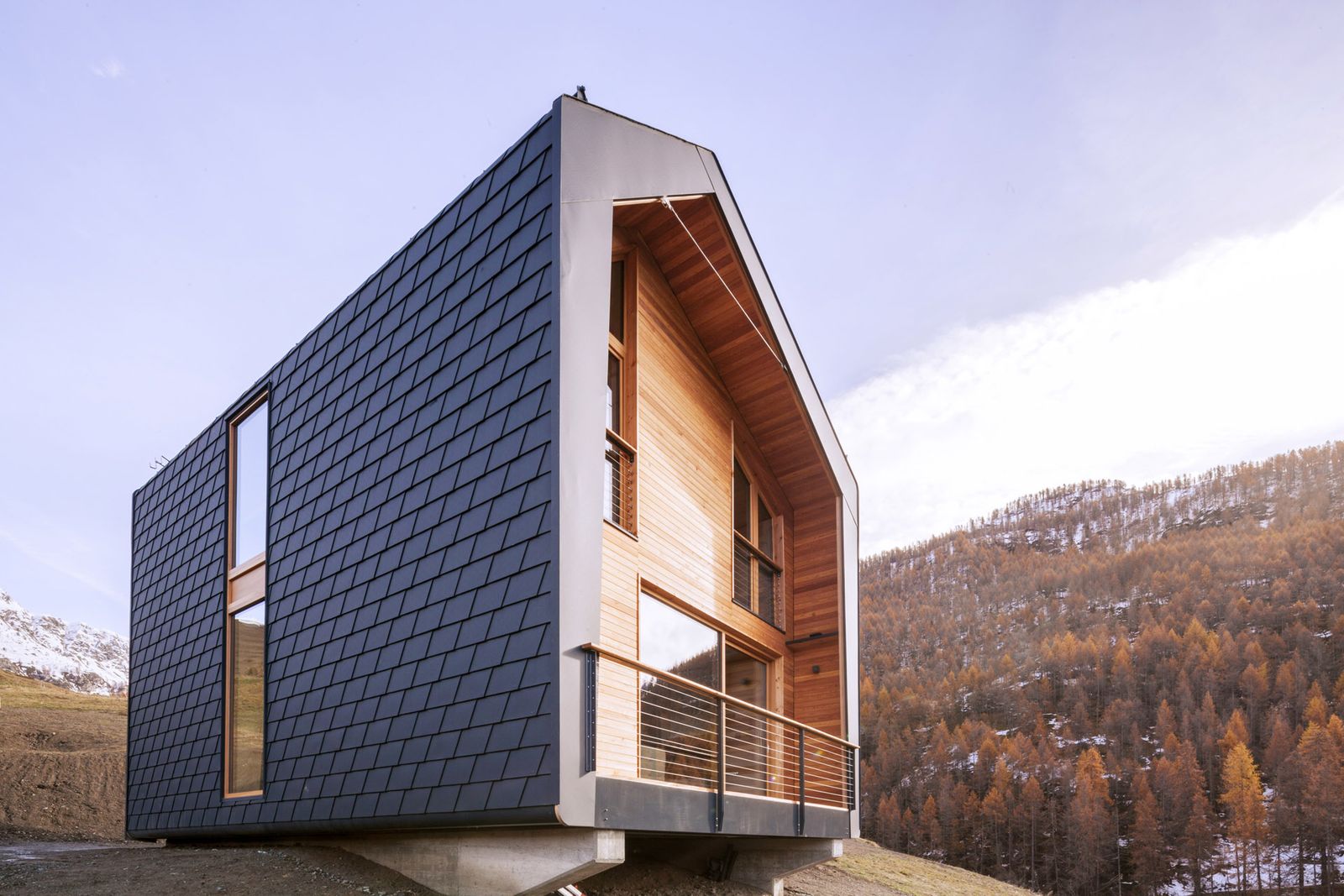 Casa a sbalzo rivestita in scandole scure con il fronte in legno e il balcone, davanti a un bosco di larici in autunno