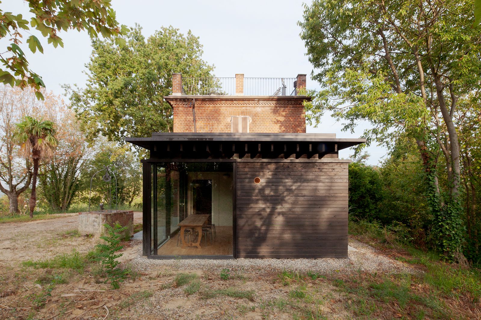 Padiglione a un piano in legno scuro con una parete vetrata e tetto sporgente, davanti a una casa storica in mattoni fra gli alberi