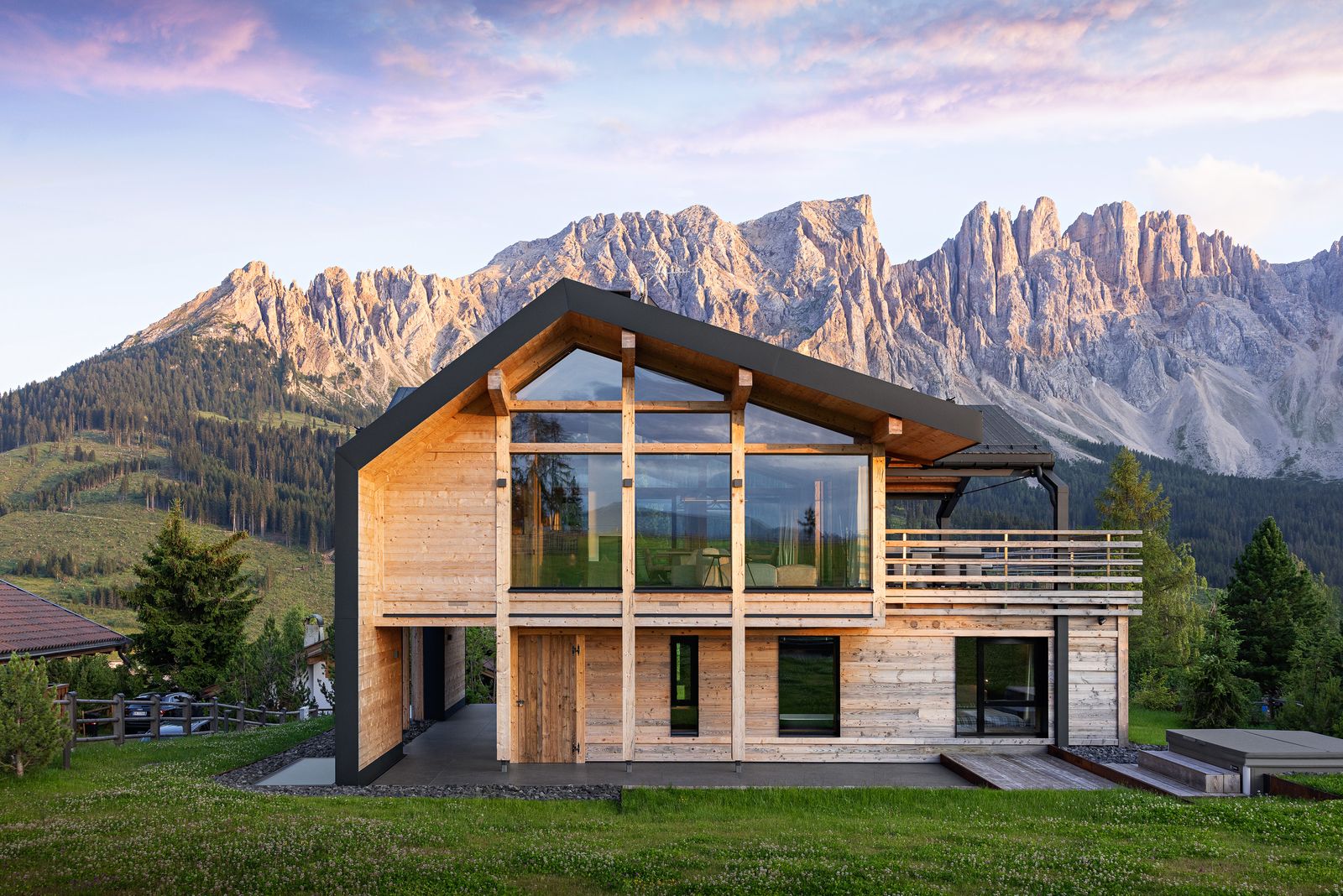 Chalet in legno chiaro con grande vetrata a capanna e balcone, su un prato davanti alle cime del Latemar all'alba