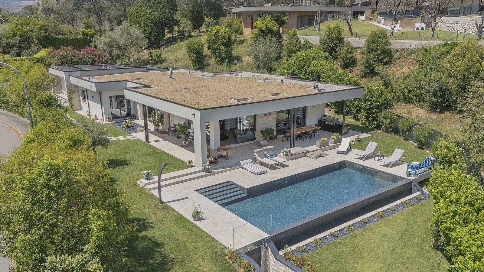 Vista dall'alto di una villa a un piano con tetto piano verde, portico, solarium e piscina a sfioro fra gli ulivi
