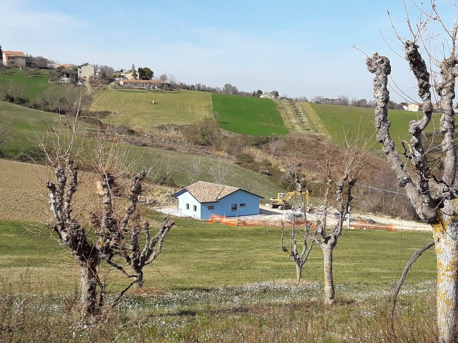 Casa in legno a un piano dipinta di azzurro, con tetto in coppi, fra le colline coltivate delle Marche