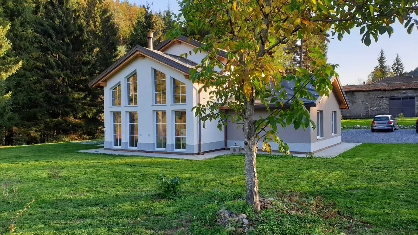 Casa in legno intonacata di bianco con grande vetrata sul frontone, prato e albero in primo piano nella luce d'autunno