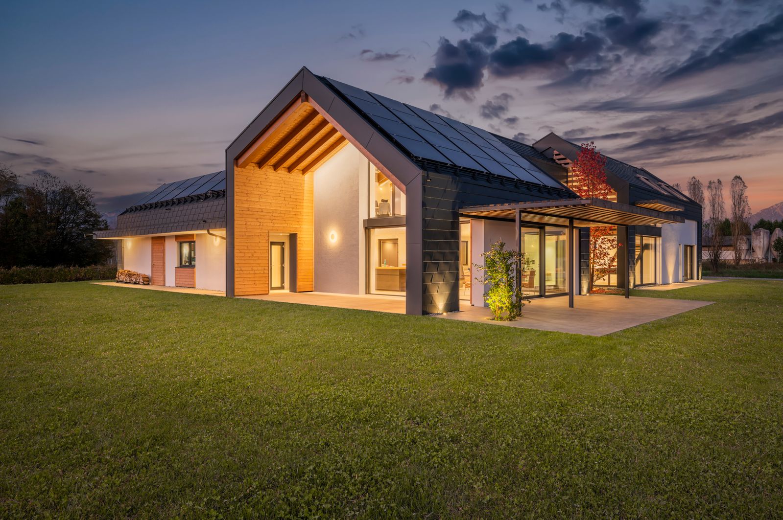 Villa unifamiliare in legno Rubner Haus al crepuscolo, con tetto a capanna fotovoltaico, pergolato e prato fra le colline del Prosecco