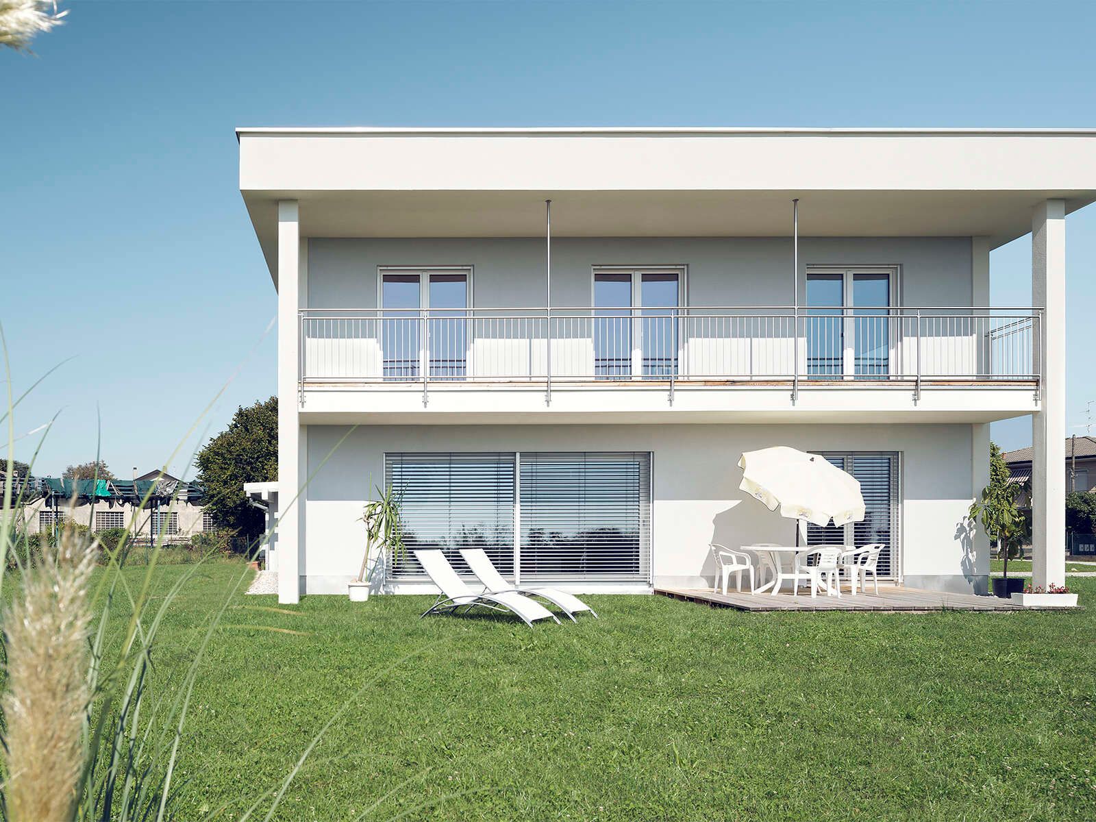 Casa in legno a due piani intonacata di bianco con tetto piano, lungo balcone e sdraio sul prato