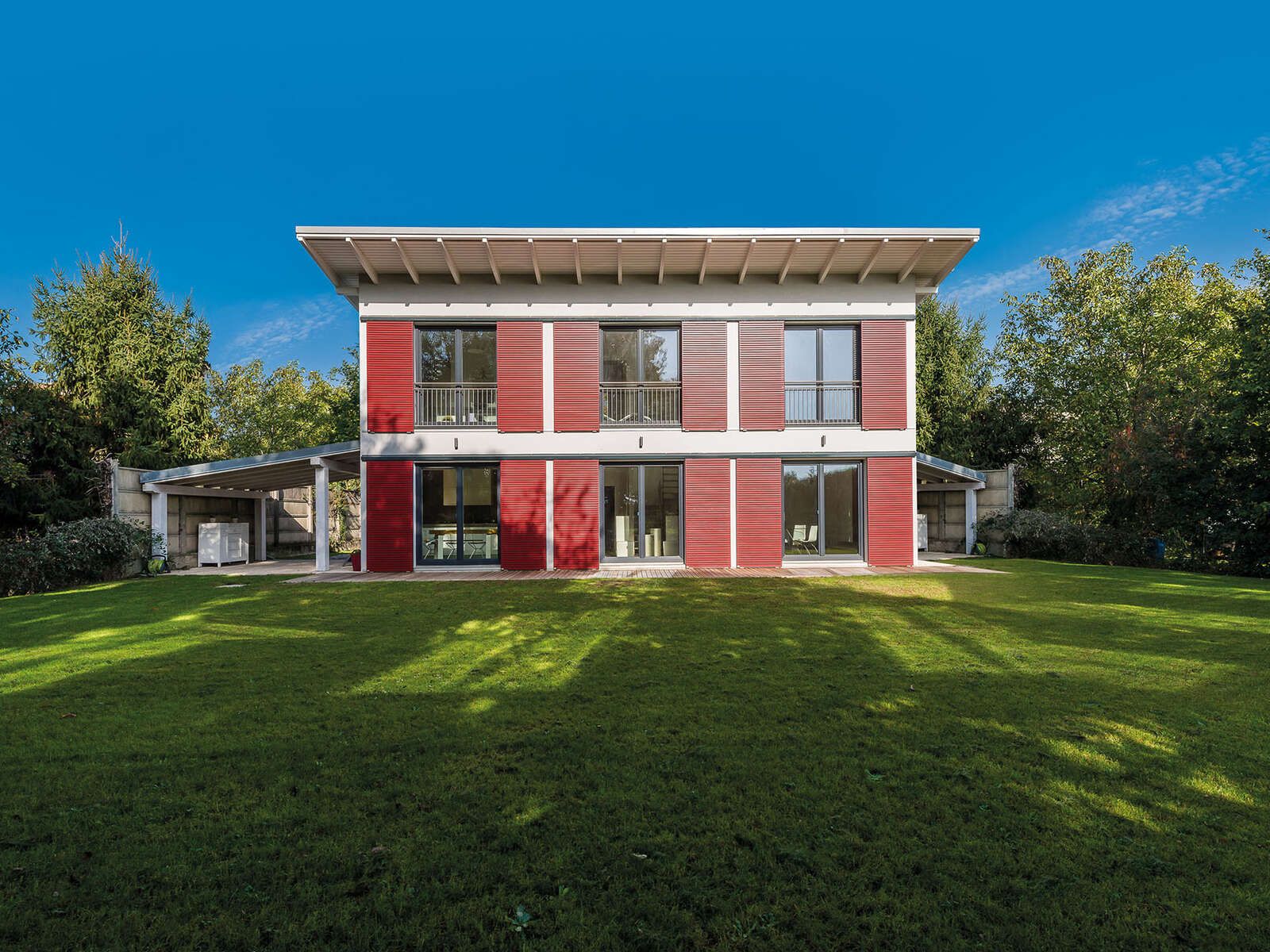 Casa in legno a due piani con facciata bianca e pannelli rossi, tetto a una falda sporgente e prato in primo piano