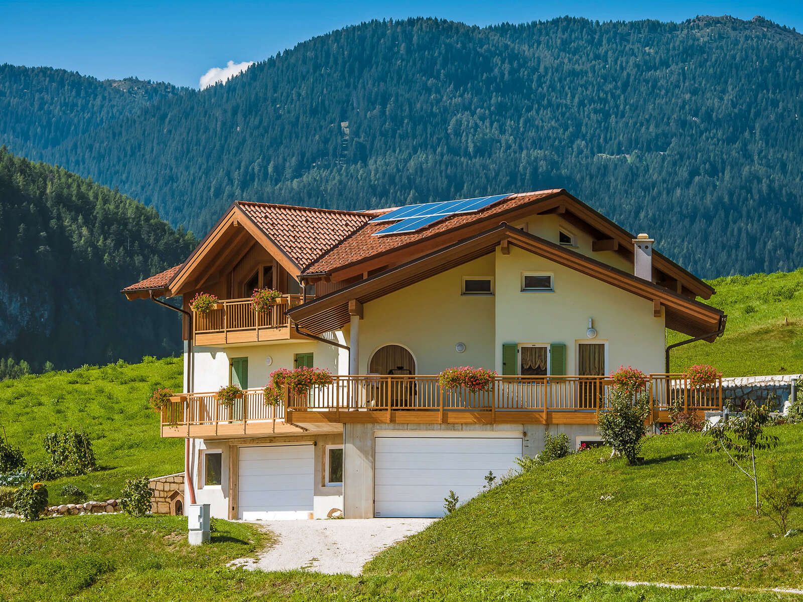 Casa in legno con tetto a falde in coppi, balconi in legno fioriti e pannelli solari, su un prato con le montagne boscose alle spalle