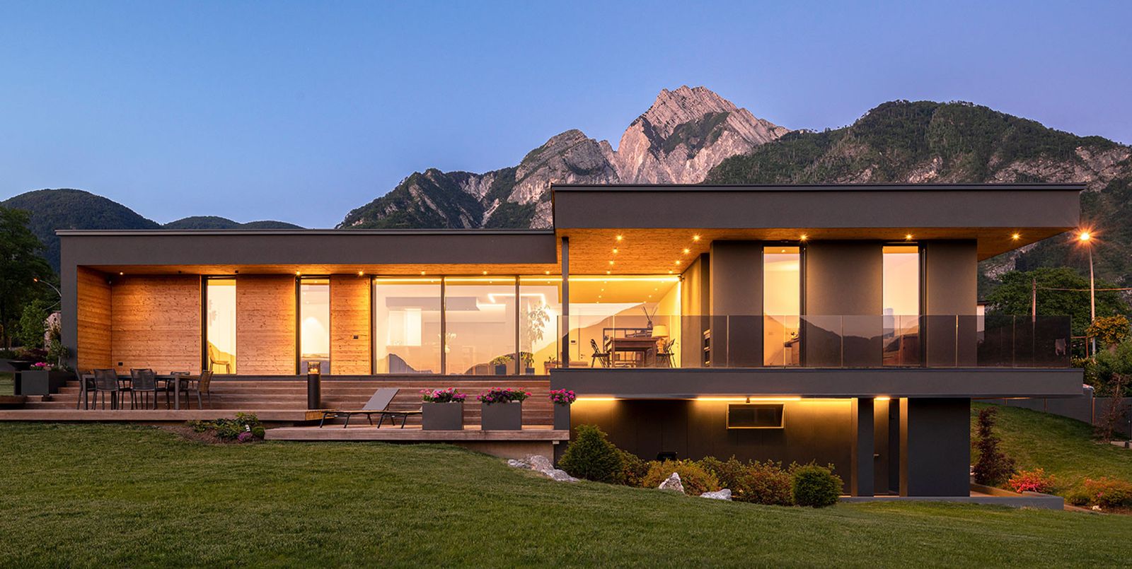 Villa Kyara di Wolf Haus al crepuscolo: volumi a tetto piano, rivestimento in legno e vetrate illuminate davanti alle montagne