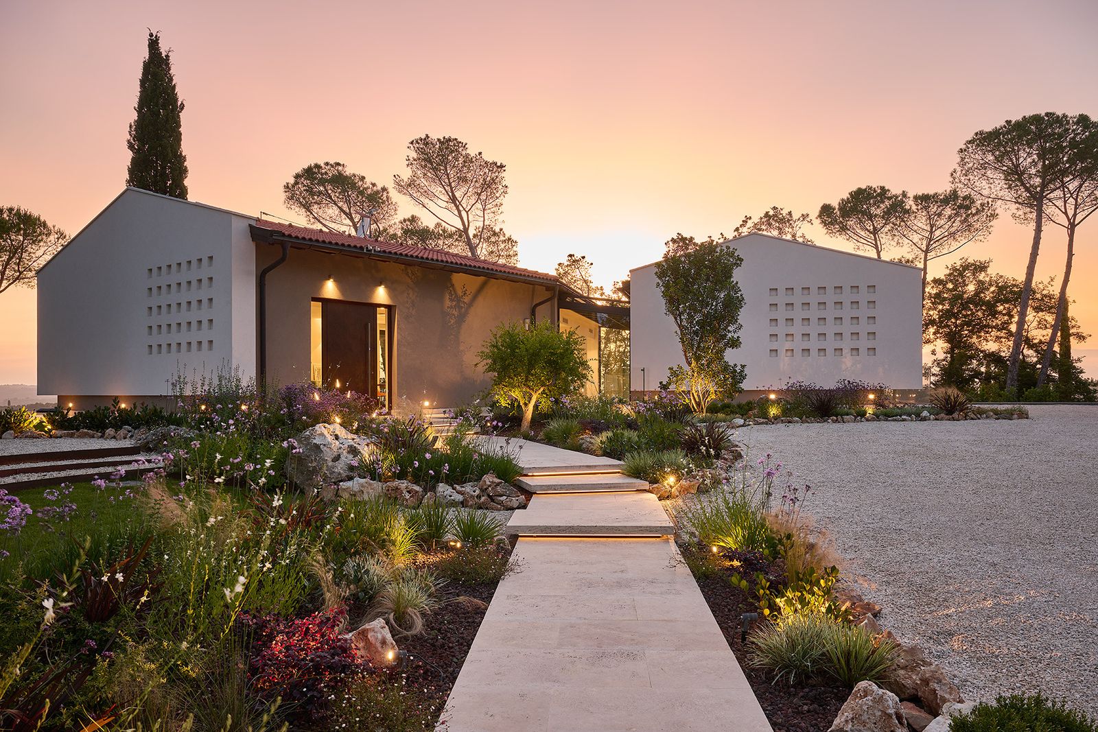 Villa Poggiolina di Wolf Haus al tramonto: due volumi bianchi a capanna, vialetto in pietra e giardino illuminato