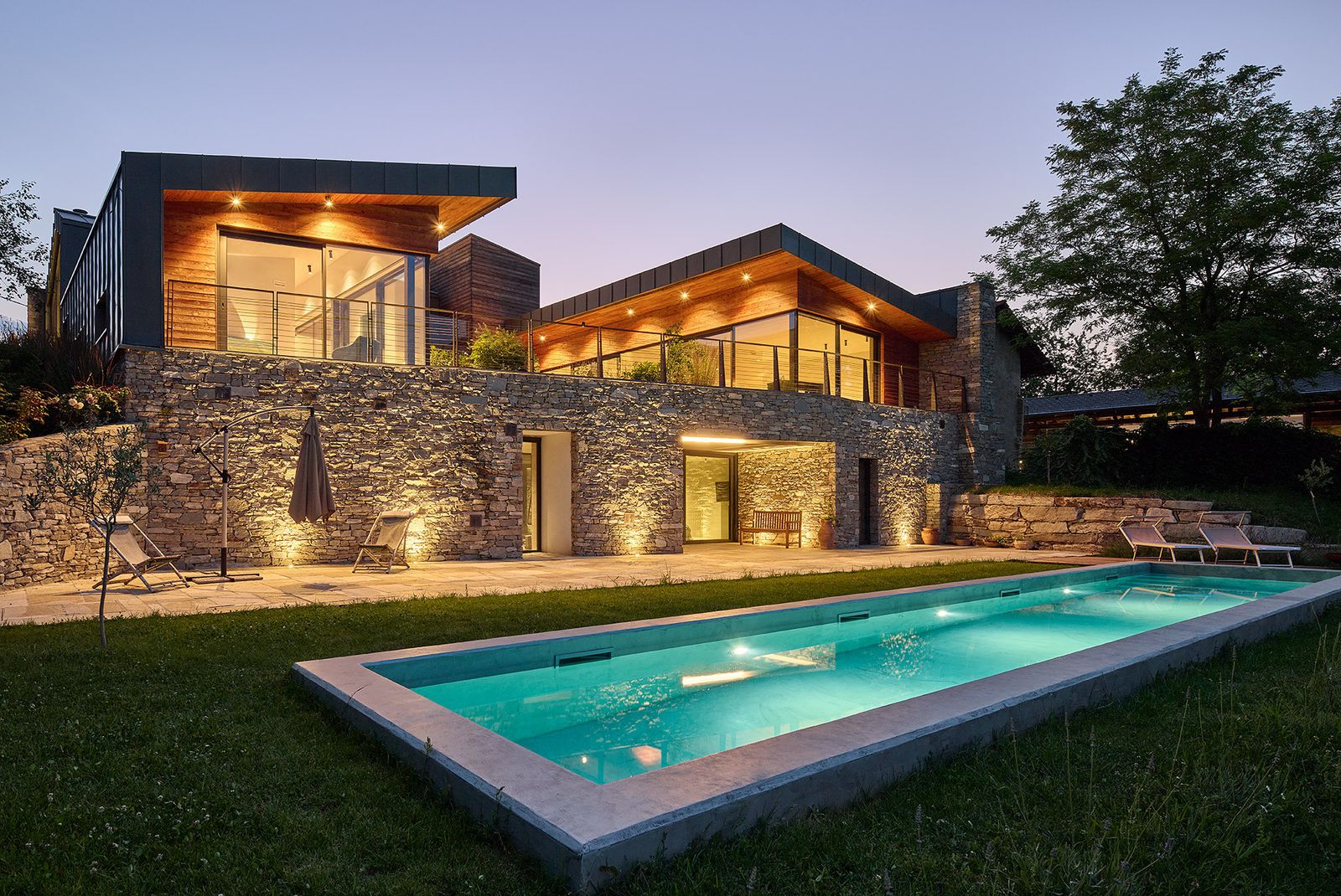 Villa Ulissys di Wolf Haus al crepuscolo: basamento in pietra, volumi rivestiti in legno e piscina illuminata nel prato